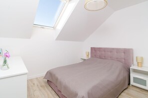 A bedroom with a bed, a desk, a nightstand, a lamp, and a window.