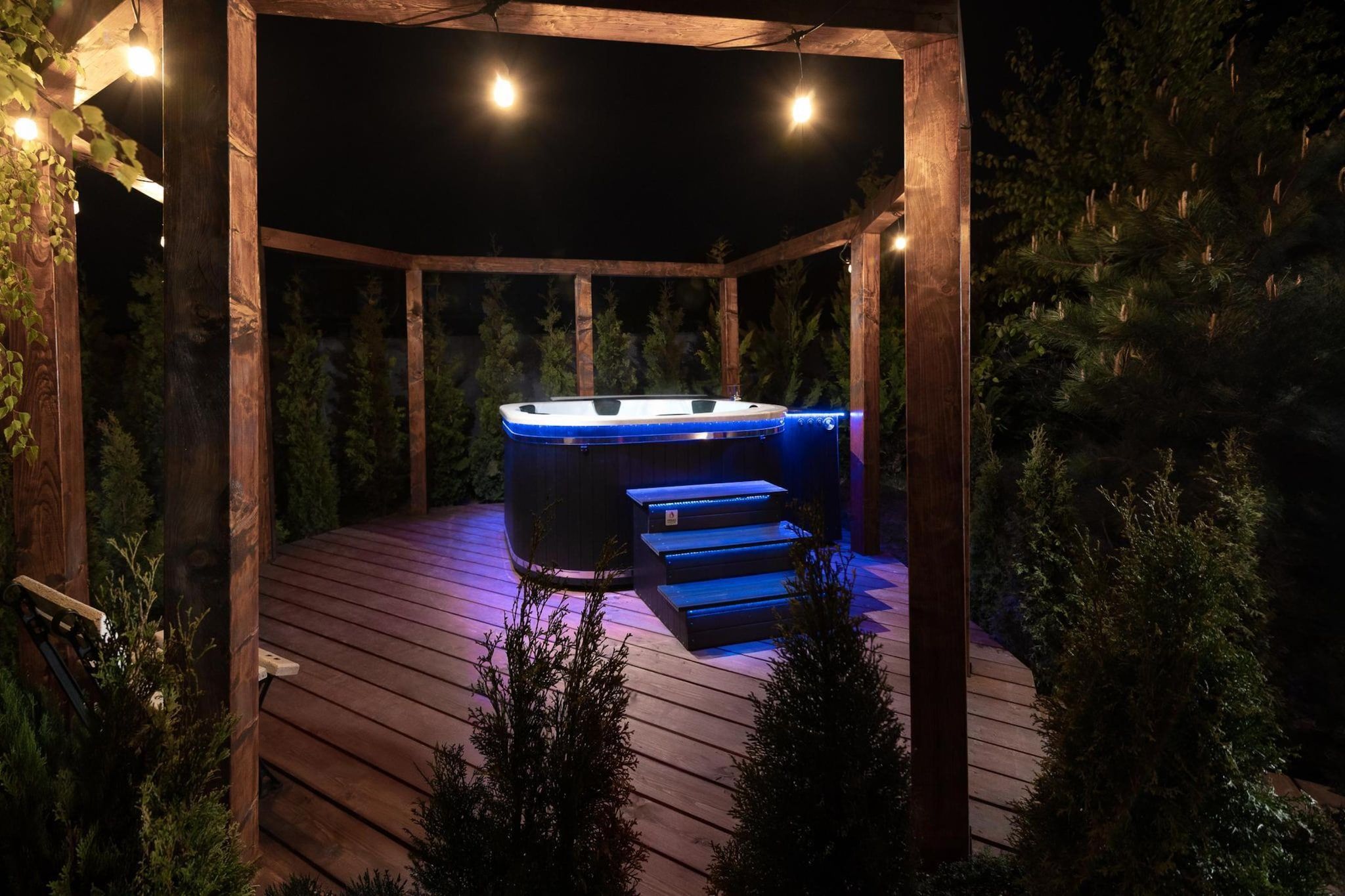 Ein Whirlpool unter einer Hollaube mit einer Holzterrasse und umgebender Bepflanzung.