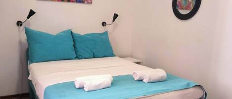 A bed with teal bedding and pillows, a white mattress, and a teal throw blanket. There is a blue rug on the floor, a wall-mounted lamp, and a decorative heart-shaped wall hanging.