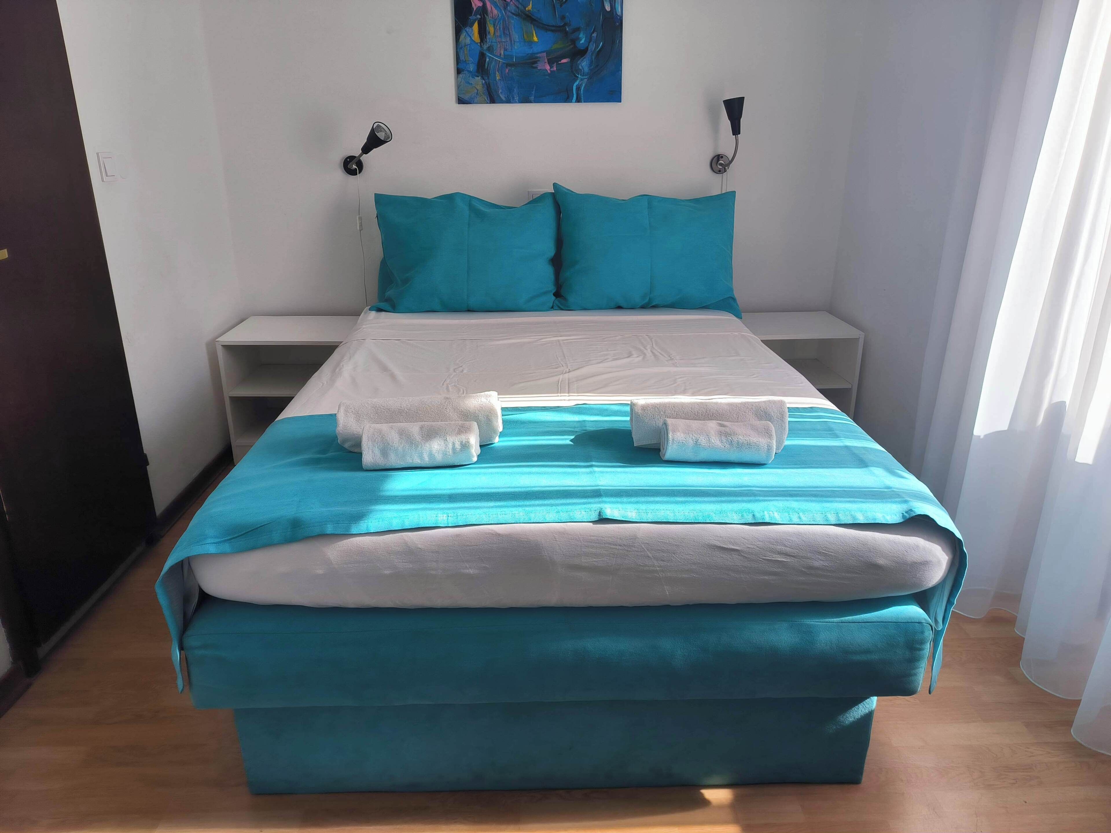 A neatly made bed with a teal bedspread and white pillows, a white nightstand with a lamp, and a painting on the wall.