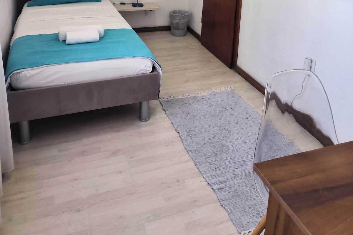 A small, clean room with a bed, a desk, a trash bin, and a door.
