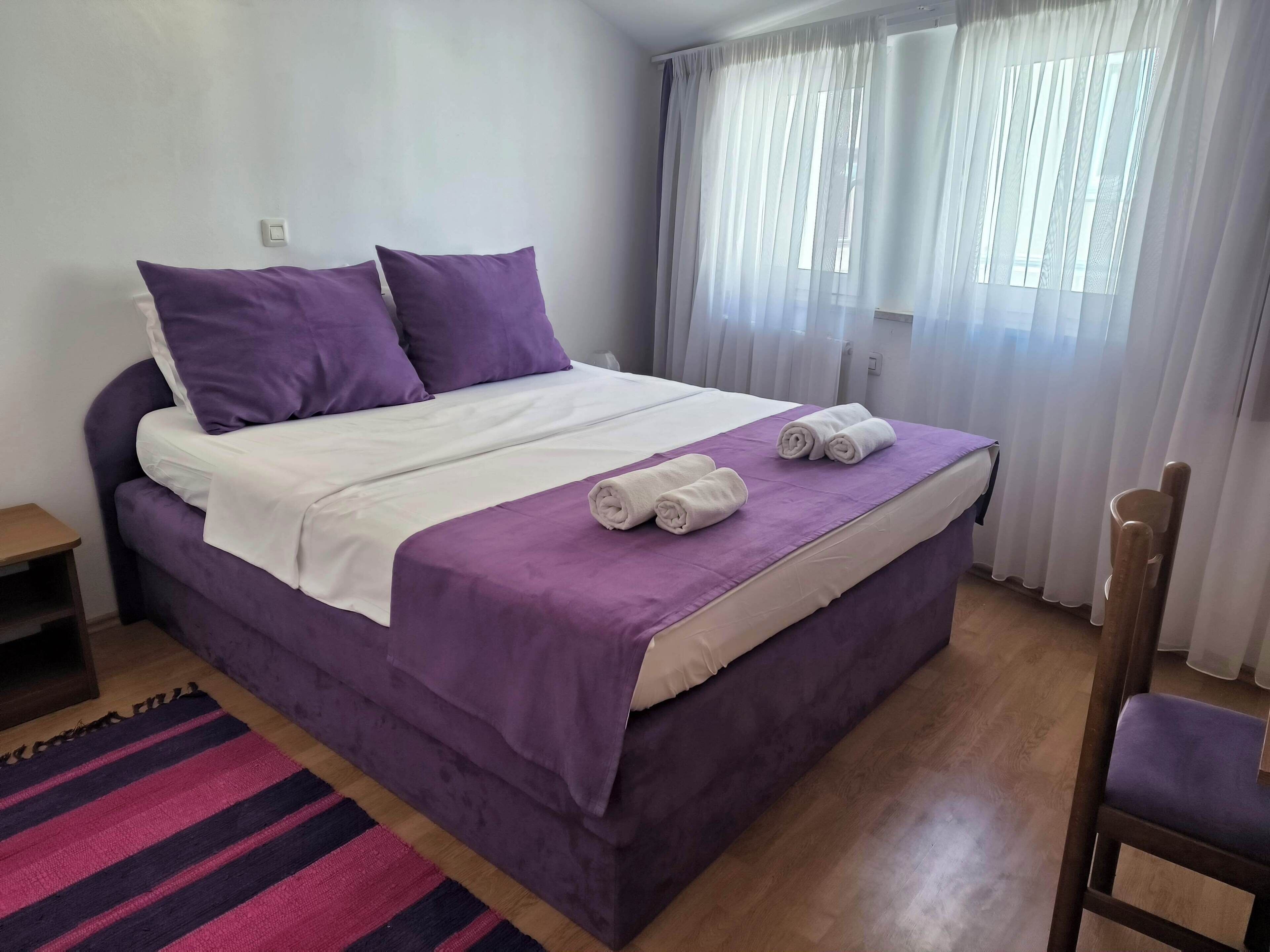 A neatly made bed with purple bedding, a purple chair, and a striped rug.
