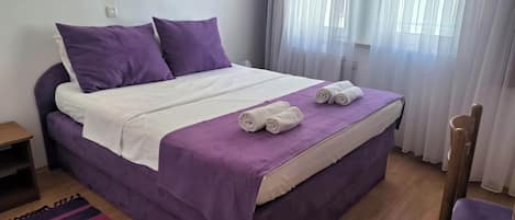 A neatly made bed with purple bedding, a purple chair, and a striped rug.