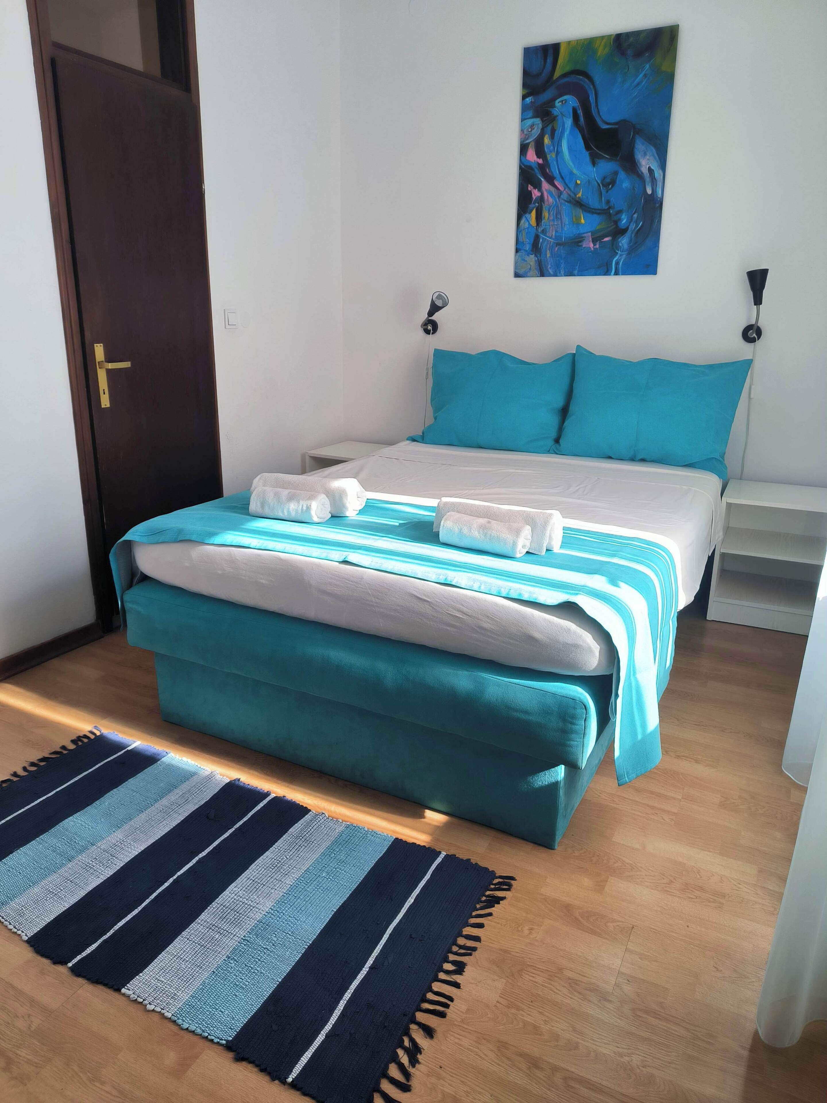 A neatly made bed with a teal and white duvet, a teal ottoman, and a striped rug on a wooden floor.