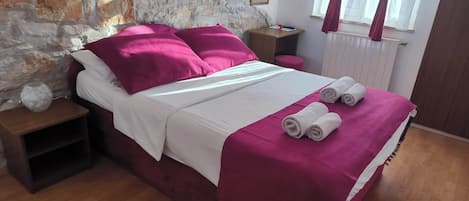 A neatly made bed with a white and pink bedspread, two pink pillows, and two rolled towels on the bed. There is a wooden nightstand with a vase and a small lamp beside the bed. The room has a stone wall, a window with red curtains, and a white radiator.