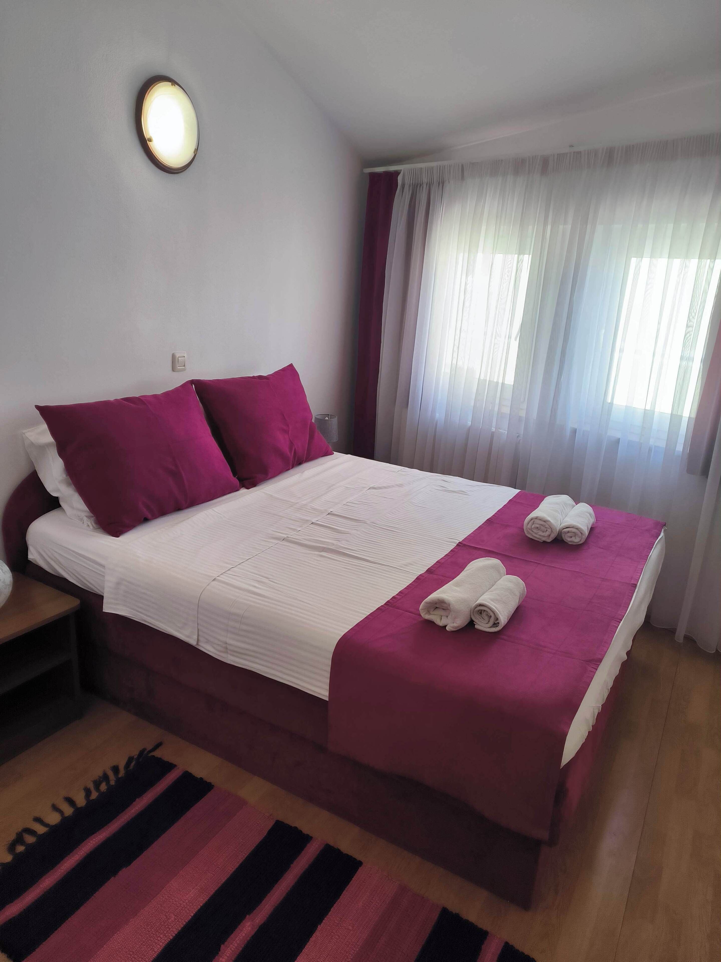 Een keurig opgemaakt bed met een wit-roze sprei, twee roze kussens en twee opgerolde handdoeken op het bed.