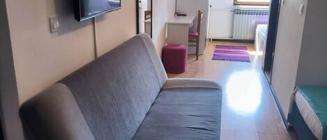 A modern living room with a grey sofa, a flat-screen TV mounted on the wall, a small table with a chair, and a view of a balcony through an open door.