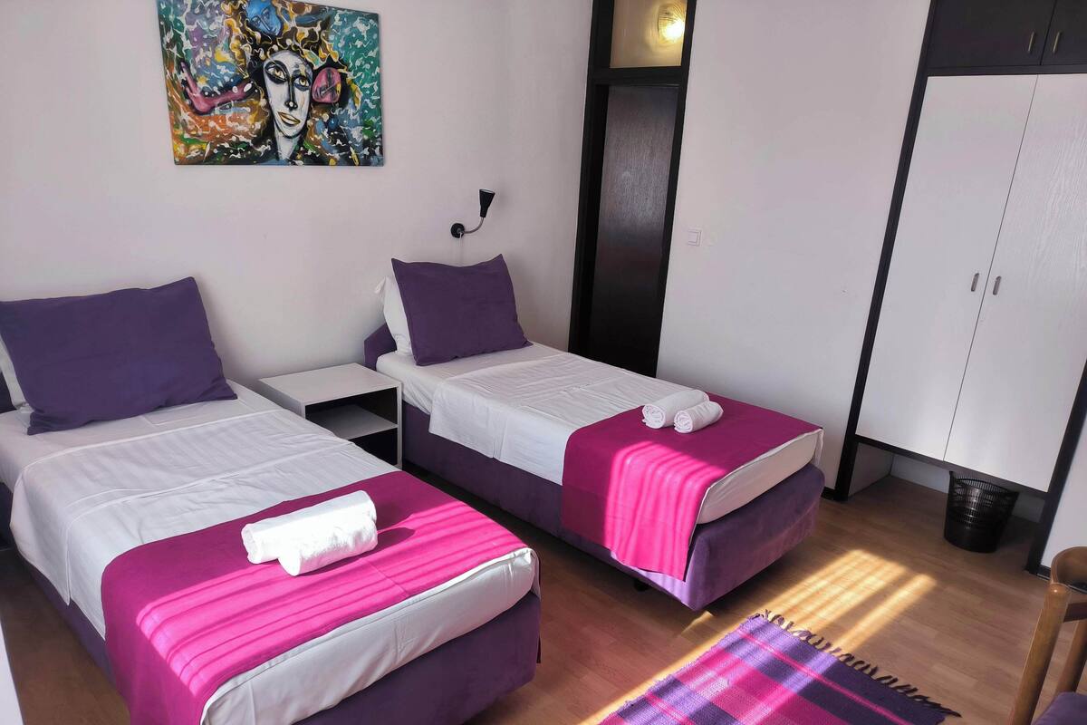 A room with two beds, each with a purple and white bedspread, a pink blanket on one bed, and a plaid rug on the floor.