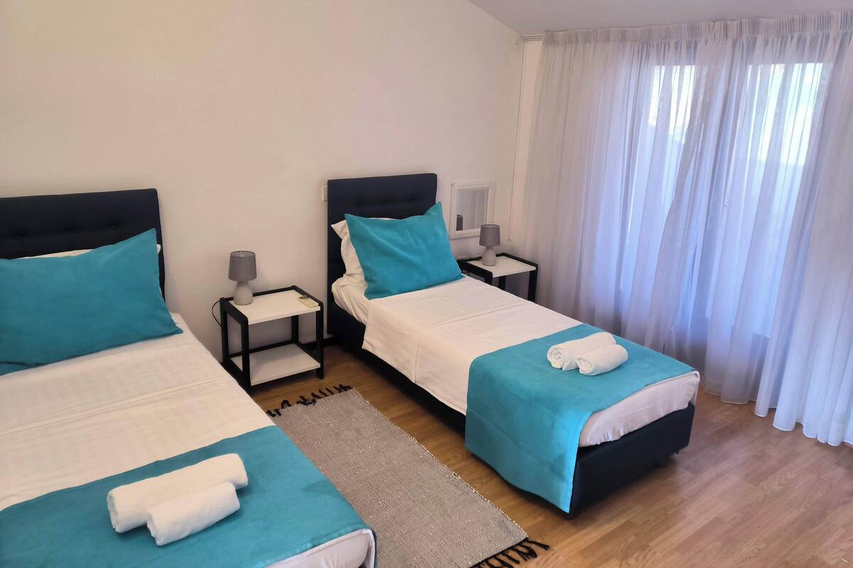 A hotel room with two beds, each with a blue bedspread and white pillows, a small bedside table with a lamp, and a large window with sheer curtains.