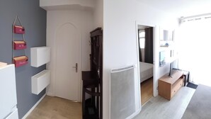 A modern hotel room with a wooden floor, a bed with a white headboard, a small wooden desk, and a chair. There is a door leading to another room and a window with curtains.
