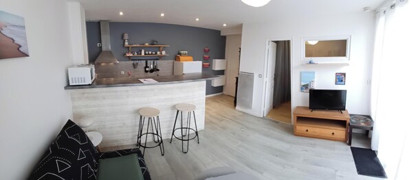 A compact living space with a kitchen area, a dining area with two stools, a sofa, a TV, and a small wooden cabinet. - Appart Biarritz Milady La Plage on foot (Biarritz)