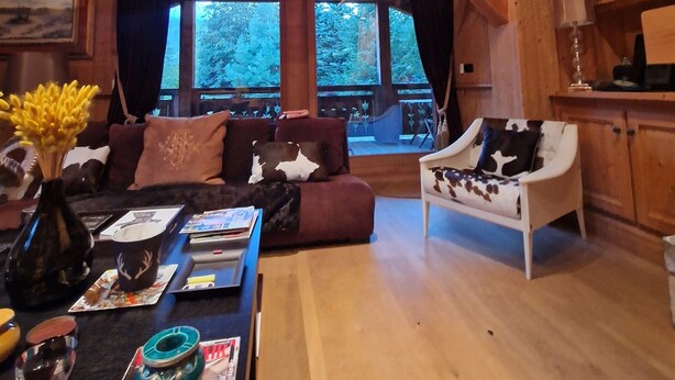 Un intérieur rustique en bois, avec un plafond haut, un coin salon confortable avec un canapé et des fauteuils, une table à manger agrémentée de magazines et d’un vase de fleurs, et de grandes fenêtres offrant une vue sur l’extérieur.