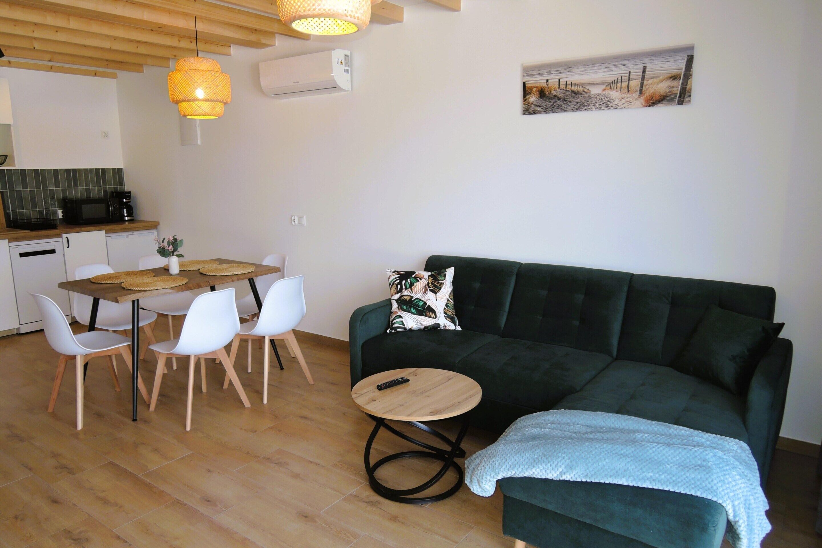Ett vardagsrum med en mörkgrön soffa, ett runt soffbord i trä, vita stolar, ett litet runt bord med en fjärrkontroll, en hängande lampa och en tavla med ett strandmotiv på väggen.