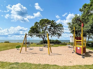 Een speeltuin met schommels en een klimstructuur, met een heldere lucht en een boom op de achtergrond.