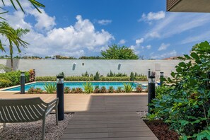 Una terraza de madera que lleva a una piscina, con un cielo azul despejado y nubes.
