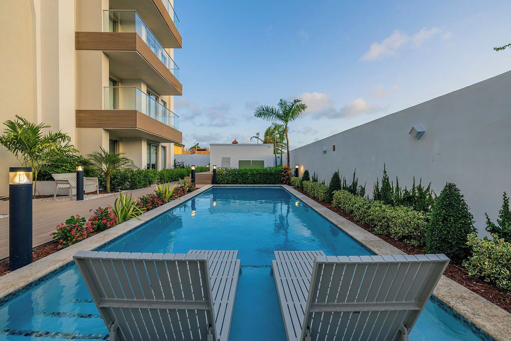 Una piscina con dos sillones de descanso, rodeada de vegetación y un edificio con balcones.
