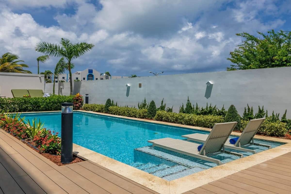 A swimming pool with lounge chairs, surrounded by greenery and a modern building in the background.