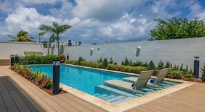 Una piscina con sillas de descanso, rodeada de vegetación y un edificio moderno al fondo.