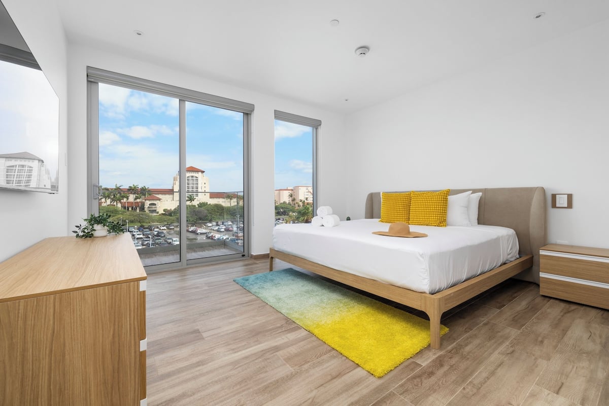 A modern bedroom with a large bed, a wooden desk, and a view of the city through large windows.