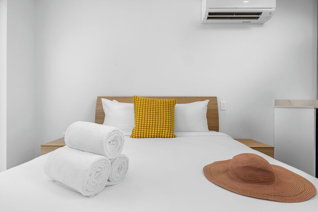 Ein Hotelzimmer mit einem Bett, weißen Bettwäsche, einem gelb gemusterten Kissen, einem braunen gewebten Hut und einer Klimaanlage.