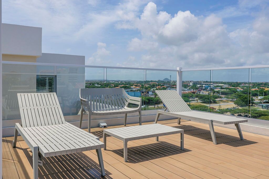 A balcony with three white lounge chairs and a small table, overlooking a cityscape with buildings and greenery.