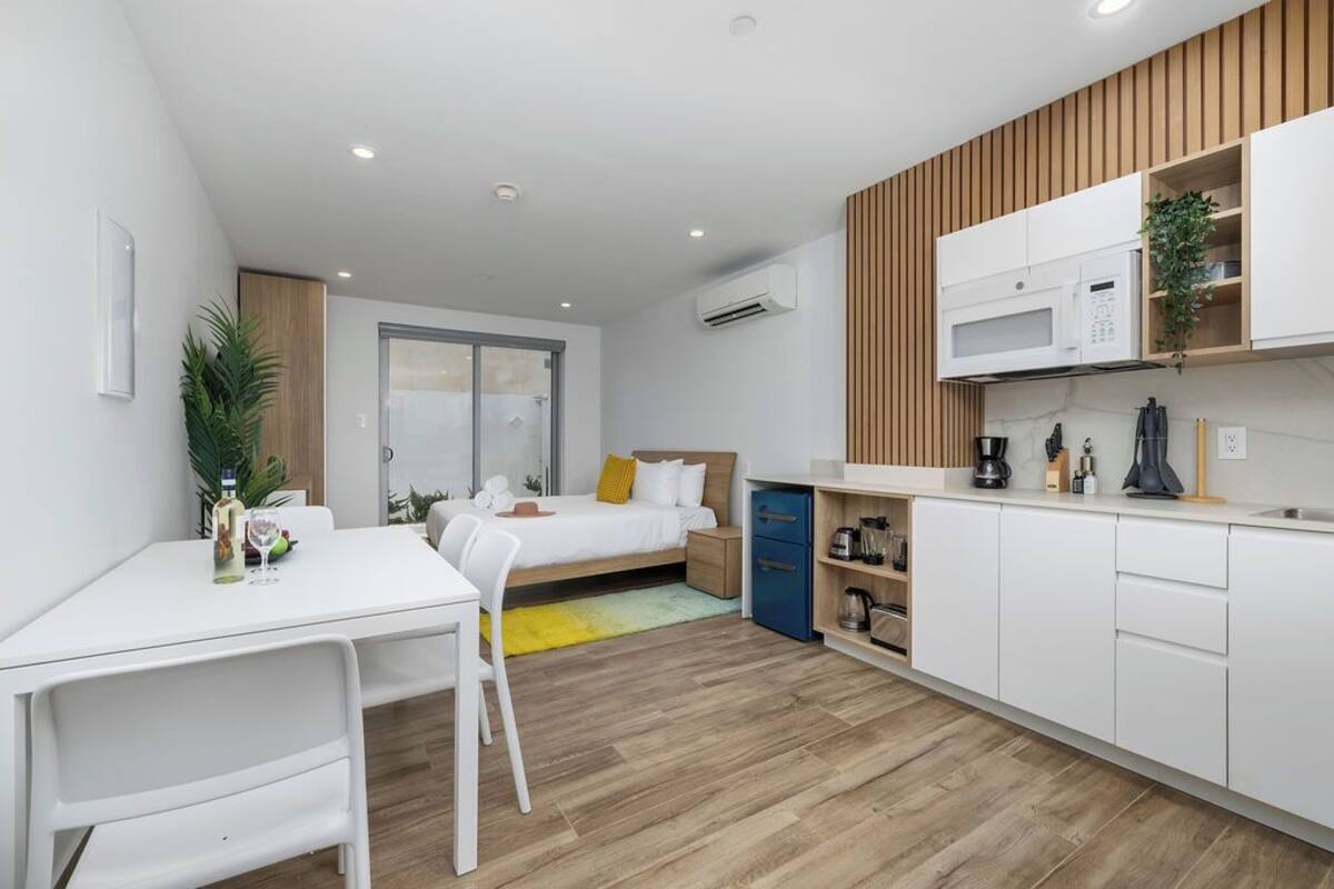 A modern, compact room with a bed, a dining table, a kitchen area, and a small seating area.