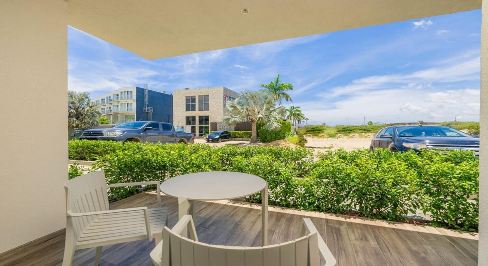 A balcony with a round table and chairs, overlooking a parking area with cars and a building in the background.