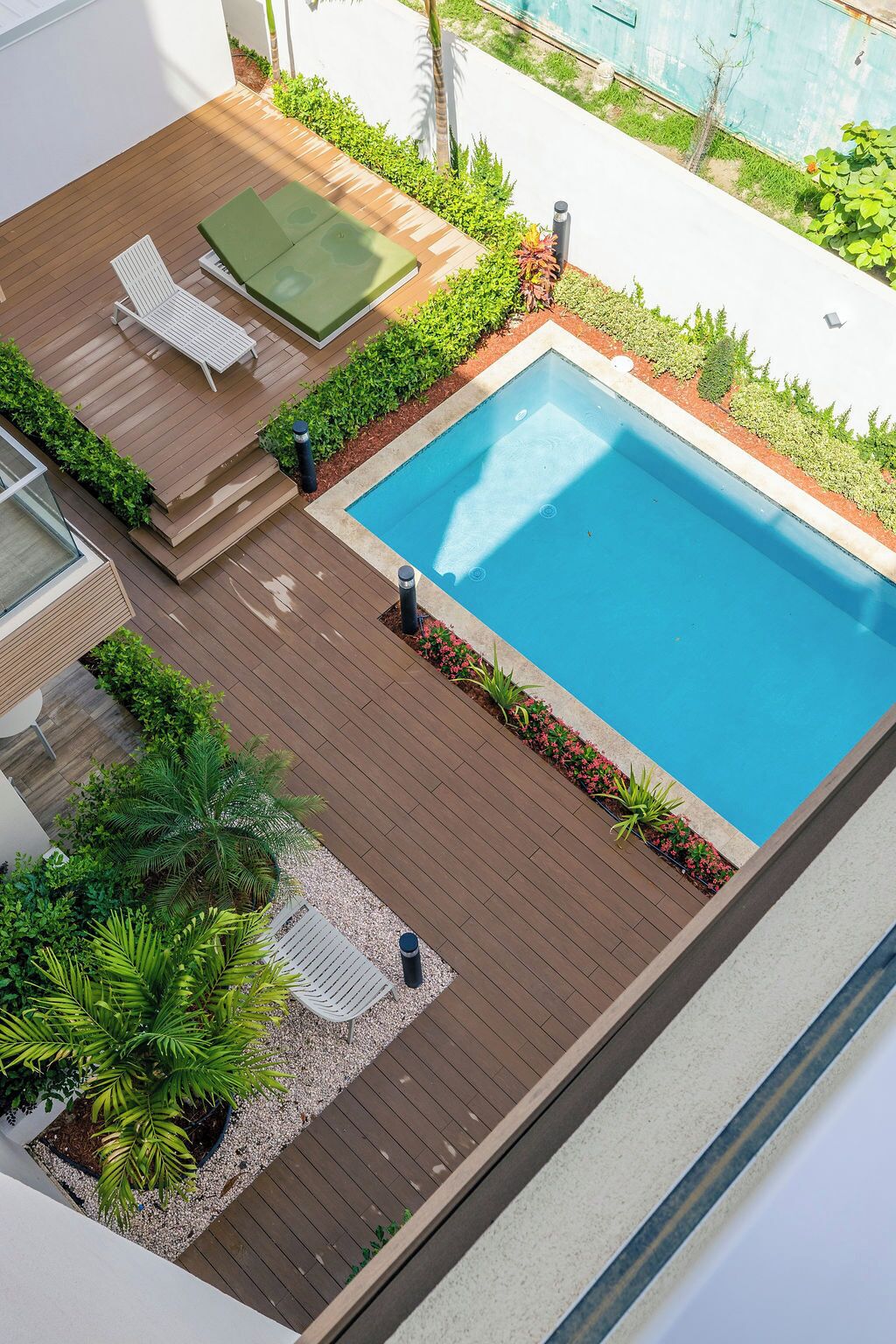 A rooftop pool area with a wooden deck, two lounge chairs, and a small garden with various plants.