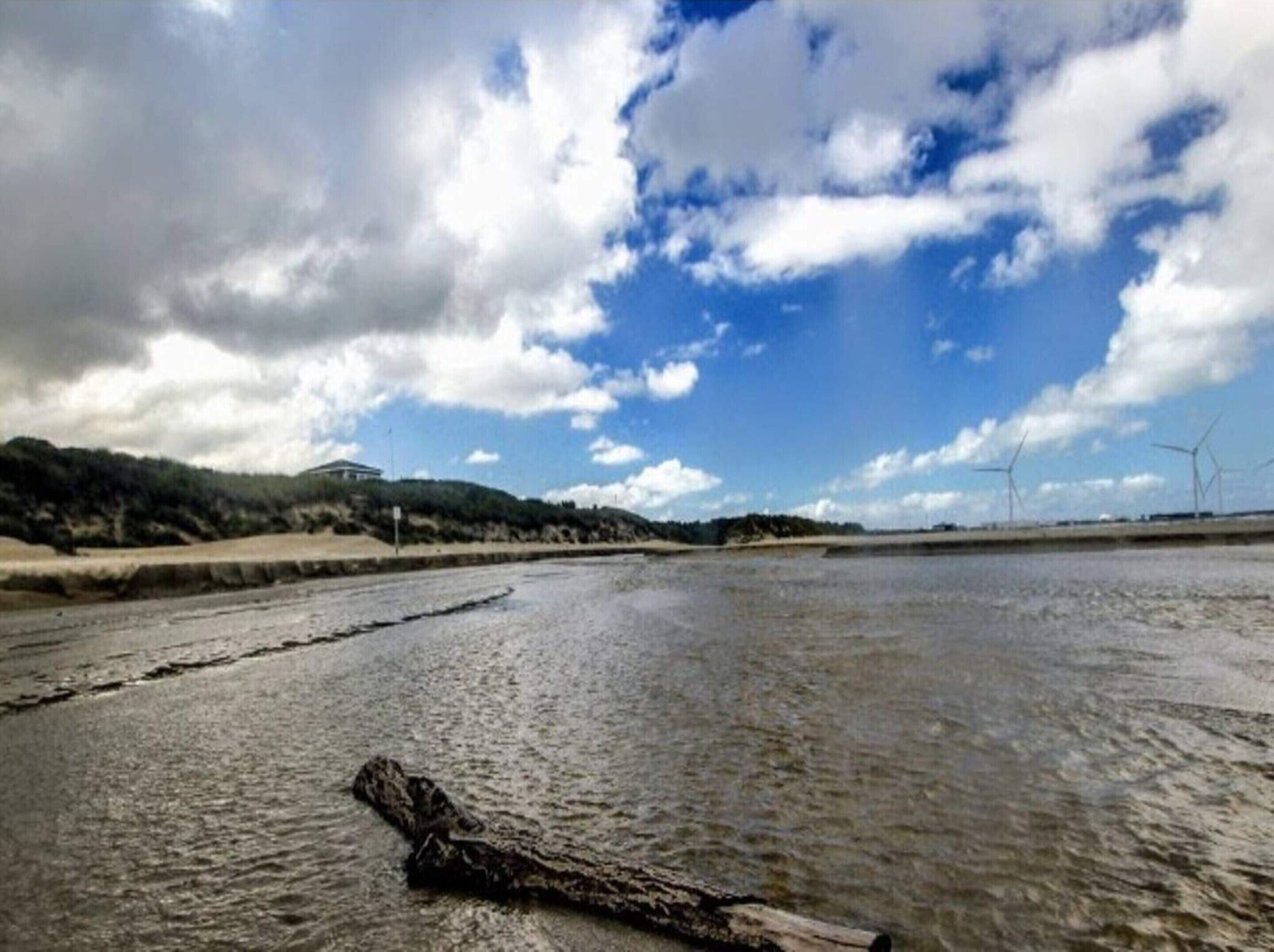 A river with a log in the foreground, a building on a hill, and wind turbines in the distance.