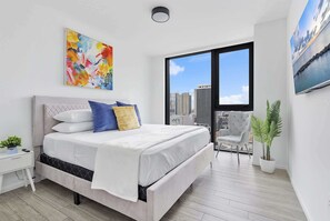 A modern bedroom with a bed, a nightstand, a chair, and a large window with a city view. - X Lounge VI-Premium Corner Residence (Miami)