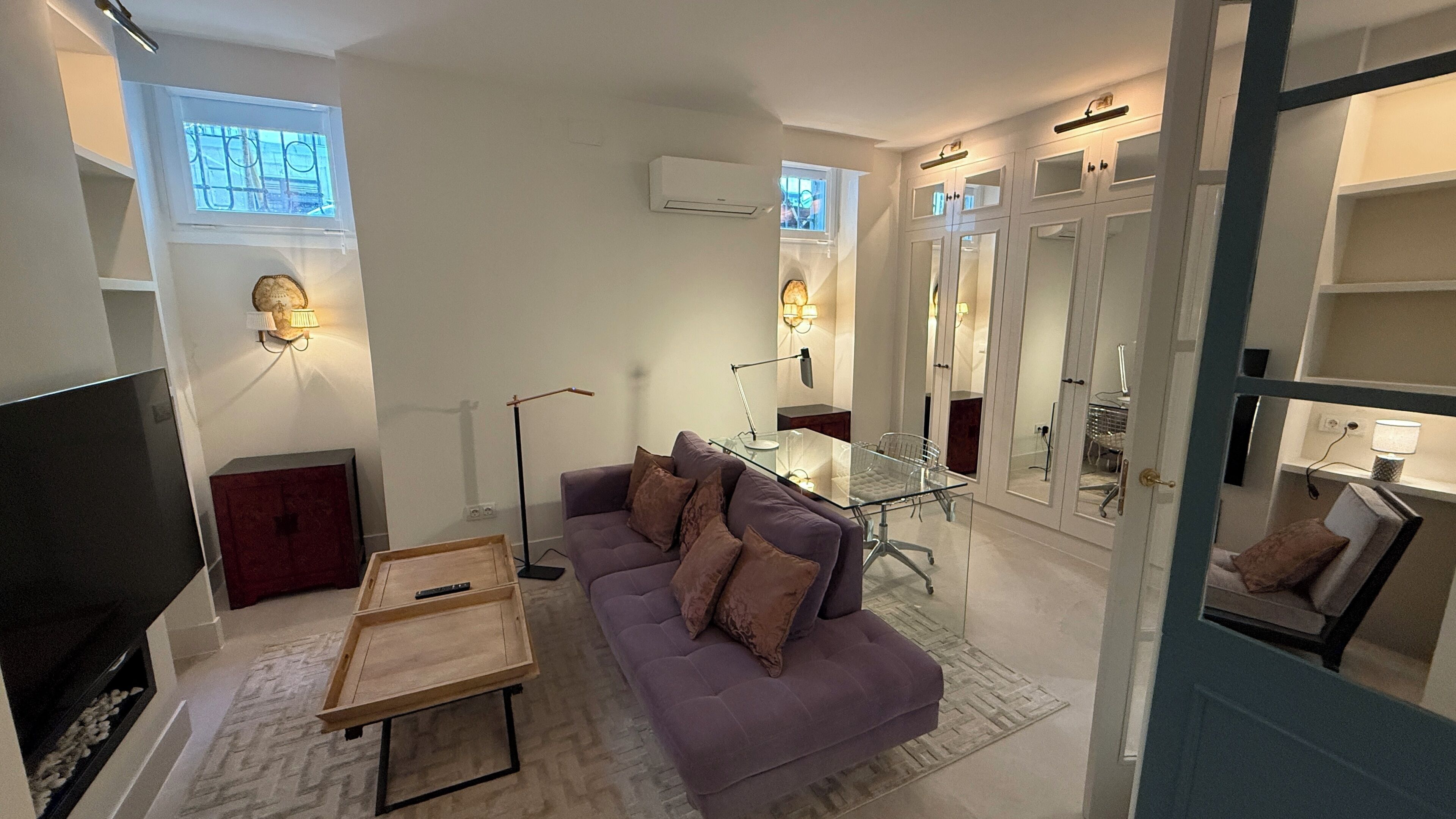 Un salon avec un canapé violet, une table basse en verre, un téléviseur à écran plat et un coin repas comprenant une table et des chaises en verre.
