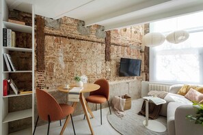 Ein modernes Wohnzimmer mit freiliegenden Ziegelwänden, einem runden Holztisch, zwei orangefarbenen Sesseln, einem weißen Sofa und einem Bücherregal voller Bücher.