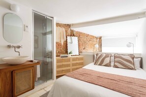 Ein Schlafzimmer mit einem großen Bett, einer Holzkommode, einem runden Spiegel, einer Glasdusche und einer Ziegelwand.
