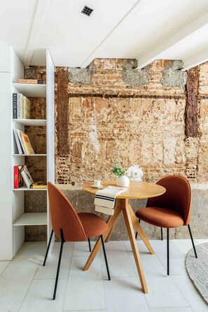 Ein Zimmer mit einer Ziegelwand, einem weißen Bücherregal, zwei orangefarbenen Stühlen, einem runden Holztisch und einer weißen Vase mit Pflanze.