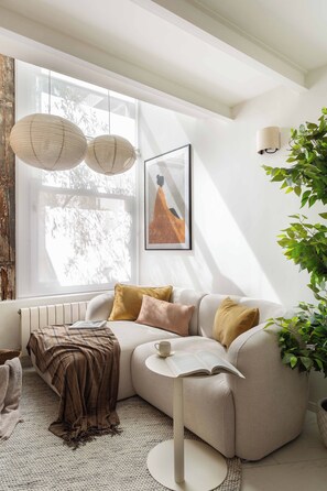 Ein modernes Wohnzimmer mit einer Eckcouch, einem runden Couchtisch, einem gerahmten Bild und einem großen Fenster mit hellem Tageslicht.
