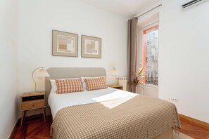 A bedroom with a bed, two bedside tables, a window with curtains, and two framed pictures on the wall.