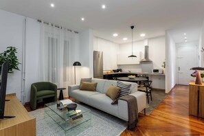 A modern living room with a sofa, a glass coffee table, a TV, and a kitchen area with a dining table and chairs.