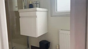 Une salle de bain avec un lavabo blanc, une cabine de douche en verre, une poubelle noire et un radiateur blanc.