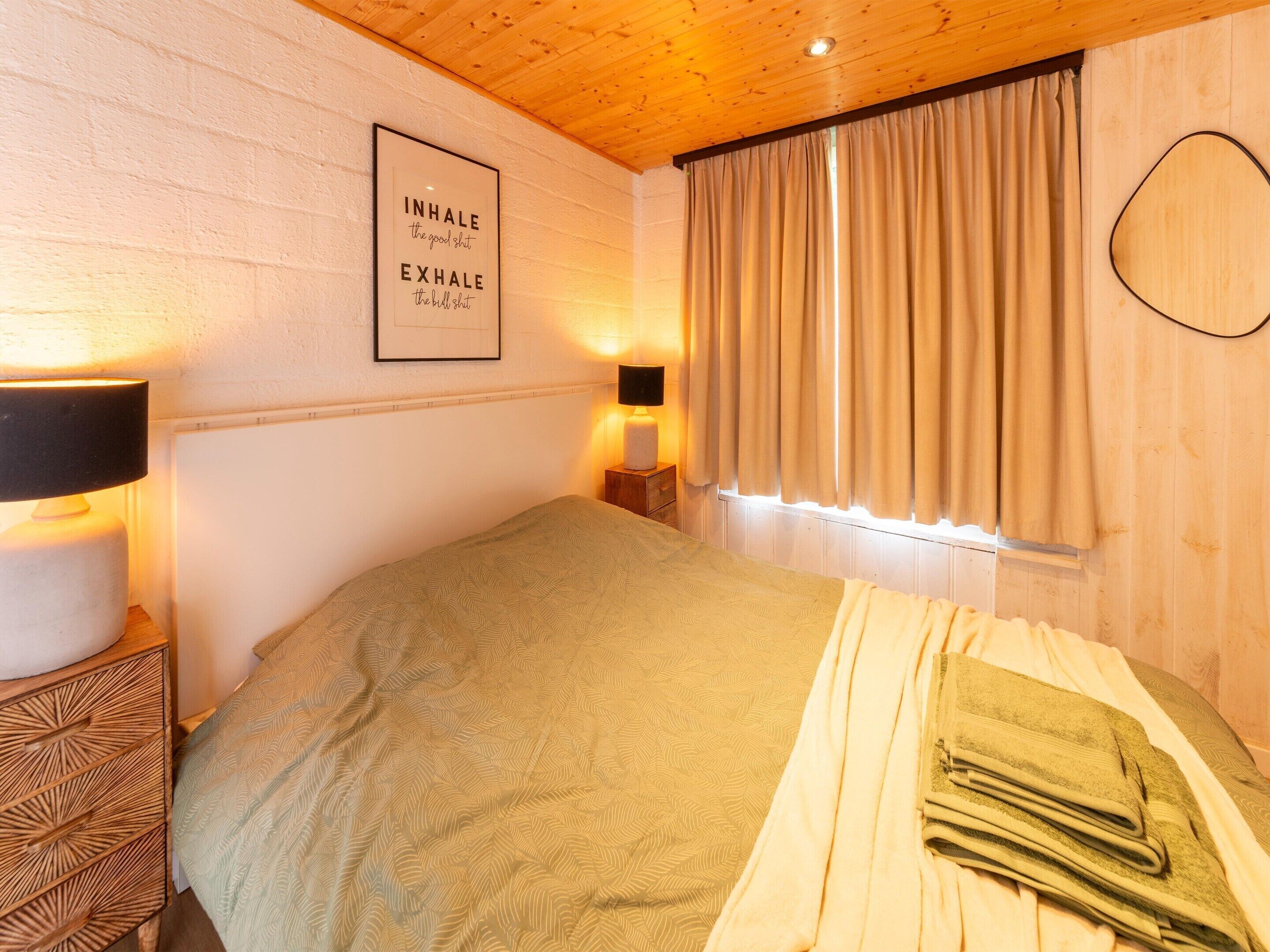 Une chambre à coucher avec un lit, une lampe de chevet, une citation encadrée accrochée au mur et une fenêtre avec des rideaux.