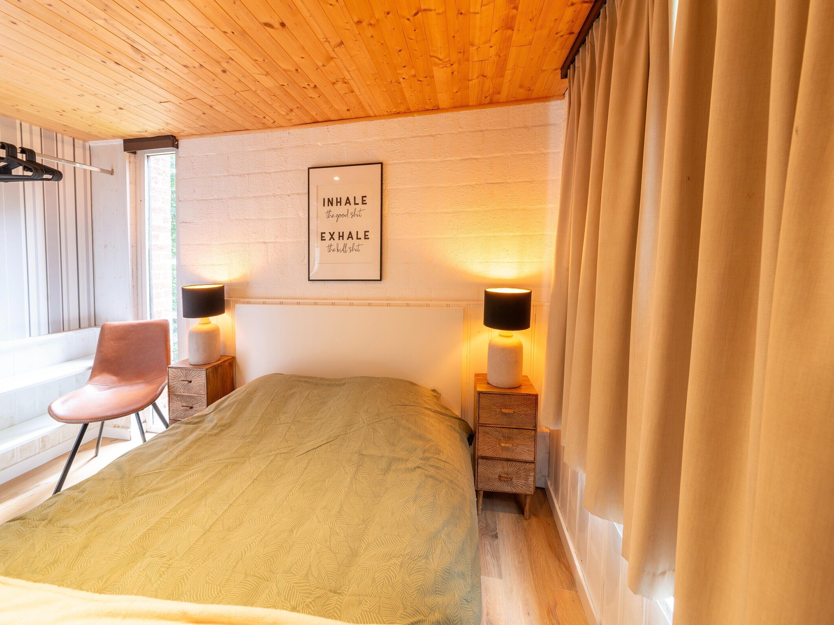 A bedroom with a bed, a chair, bedside lamps, a framed quote on the wall, and a window with curtains.