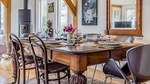 Een eethoek met een grote houten tafel, stoelen en een zwarte kookplaat. Er hangen schilderijen aan de muren en boven de tafel is een spiegel.