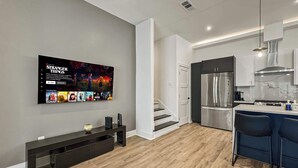 A modern living room with a flat-screen TV mounted on the wall, a dark wooden bench, and a kitchen area with a stainless steel refrigerator and a bar counter.