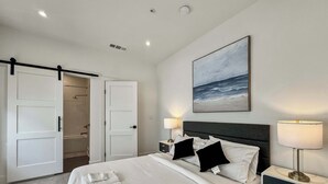 A hotel room with a bed, two bedside tables with lamps, a painting of the ocean, and a sliding door leading to a bathroom.