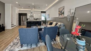 A modern living room with a dining area, a kitchen with stainless steel appliances, and a staircase leading to another level.