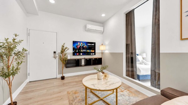 A modern living room with a flat-screen TV, a round coffee table, and a large window with curtains.