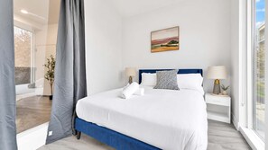 A hotel room with a large bed, two bedside tables with lamps, a window with a view, and a painting on the wall. - Posh 3BR Flat by Liberty Bell | Hosts Group of 6 (Philadelphia)