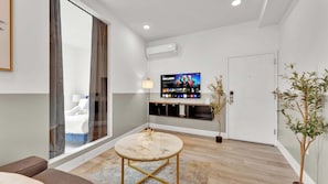 A modern living room with a flat-screen TV, a marble coffee table, and a large window with curtains.