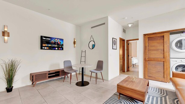 Une chambre d’hôtel moderne équipée d’un téléviseur à écran plat, d’une table de salle à manger ronde, de deux chaises, d’un banc, d’une plante en pot et d’une armoire en bois contenant une machine à laver.