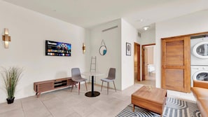 Une chambre d’hôtel moderne équipée d’un téléviseur à écran plat, d’une table de salle à manger ronde, de deux chaises, d’un banc, d’une plante en pot et d’une armoire en bois contenant une machine à laver.