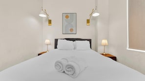 Een net opgemaakt bed met witte lakens en opgerolde handdoeken, twee nachtlampjes, een kunstwerk met abstracte afbeelding en een raam met een zonnescherm.